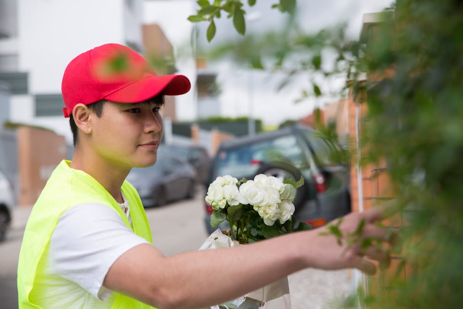 A young male flower delivery person wearing a red cap and a high-visibility yellow vest hands over a bouquet of fresh white hydrangeas to a customer outdoors on a residential street. The bouquet is wr