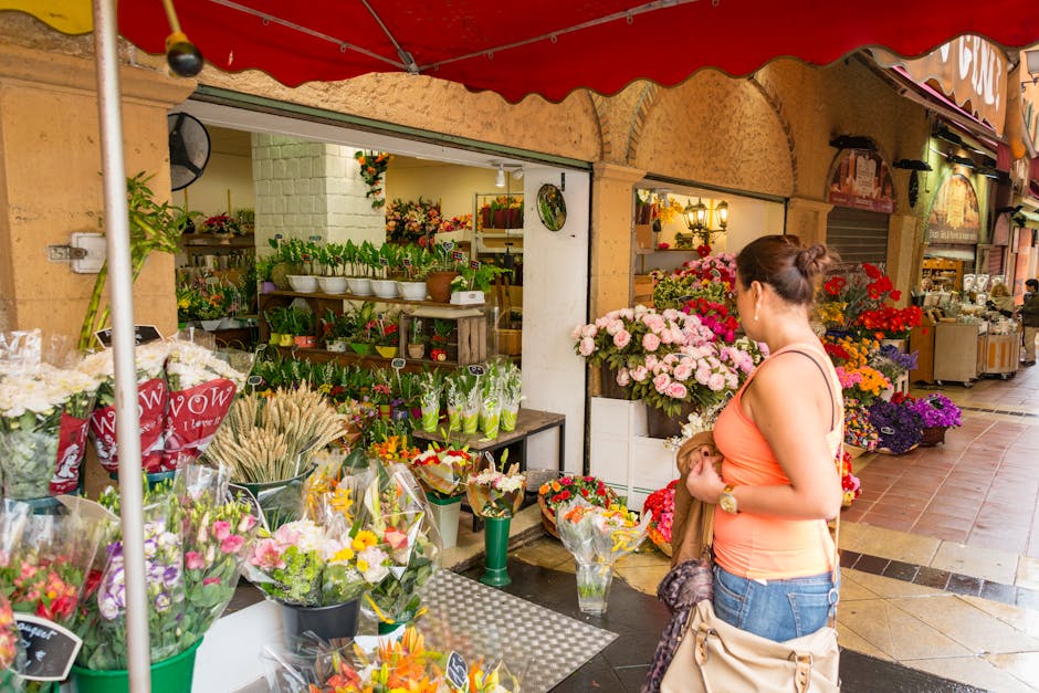 A woman wearing an orange sleeveless top and blue jeans is holding a small bouquet of flowers while standing outside a florist shop on a busy street. The shop displays a vibrant array of floral arrang