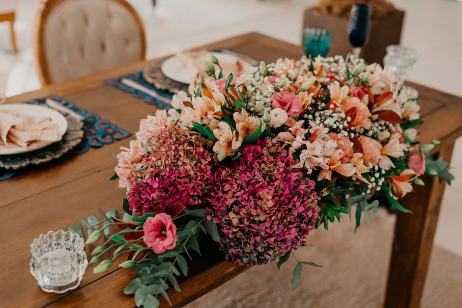 A large, vibrant floral arrangement placed on a wooden dining table features a combination of pink and peach-colored flowers, including hydrangeas, alstroemerias, and baby's breath, accented with gree