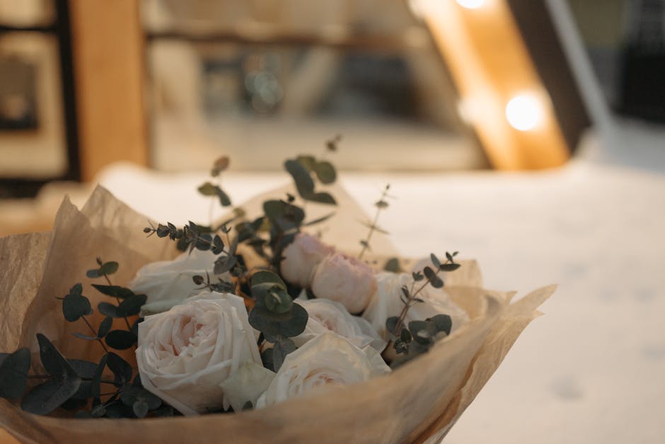 A bouquet of pale pink and cream roses arranged with sprigs of dark green eucalyptus leaves, wrapped in light brown paper. The bouquet is positioned on a surface with a blurred background featuring wa