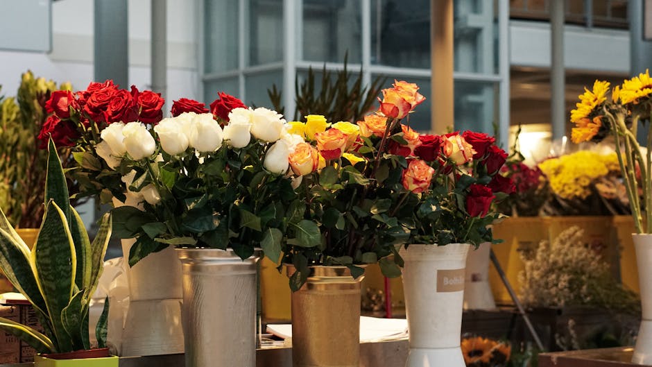 A display of fresh floral arrangements featuring red, white, and yellow roses, arranged in metal and ceramic vases. The bouquets are placed on a table indoors, with some flowers still wrapped in flora